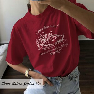 May include: Red short-sleeved t-shirt with a white graphic of two swans and the text "I know love is real because I exist and I'm full of it".