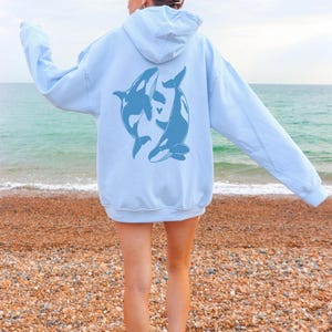 May include: A light blue hoodie with a graphic of two orcas on the back. The orcas are facing each other and appear to be swimming in a circle. The hoodie is worn by a person standing on a beach.