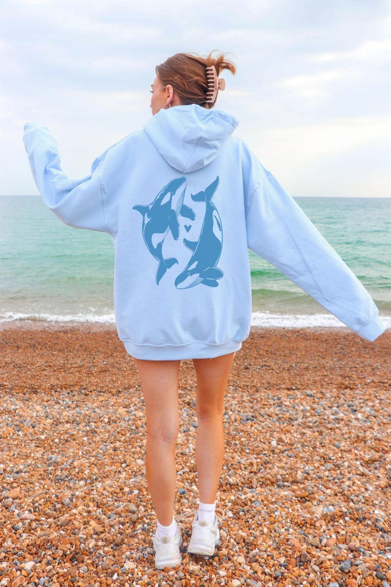 May include: A light blue hoodie with a graphic of two orcas on the back. The orcas are facing each other and appear to be swimming in a circle. The hoodie is worn by a person standing on a beach.