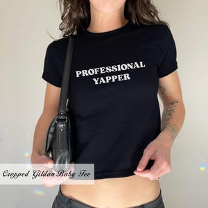 May include: A black cropped t-shirt with the text "PROFESSIONAL YAPPER" in white lettering. The shirt is being worn by a person with a black shoulder bag.