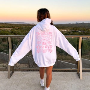 May include: White hoodie with a pink graphic print on the back. The graphic features a variety of images including teacups, flowers, and the text "Painted your heart".