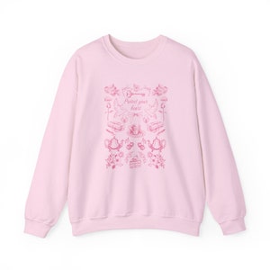 May include: A pink sweatshirt with a pink graphic print of a key, birds, teacups, and flowers. The text "Protect your heart" is printed on the sweatshirt.