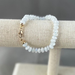 May include: A white gemstone bracelet with a gold clasp. The bracelet is made of small, round beads and is displayed on a fabric-covered stand.