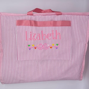 May include: Pink and white striped fabric tote bag with a pink handle and a pink pocket. The pocket has the name "Lizbeth" embroidered on it in pink thread, along with a pink bow and a string of colorful hearts.