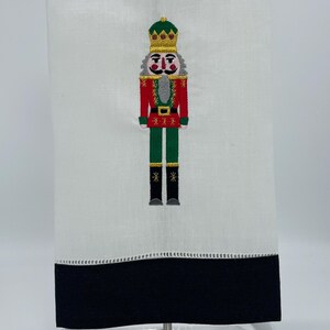 May include: White kitchen towel with a black border and a red, green, and gold embroidered nutcracker design.