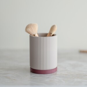 May include: A grey ribbed ceramic makeup brush holder with a burgundy base. Two makeup brushes are inside the holder.