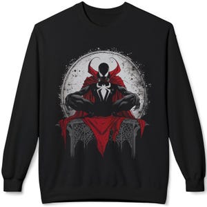 May include: Black sweatshirt featuring a comic book illustration of a character with a red cape and a white spider emblem. The character is seated in front of a gray moon with a distressed texture. The design is centered on the front of the sweatshirt.