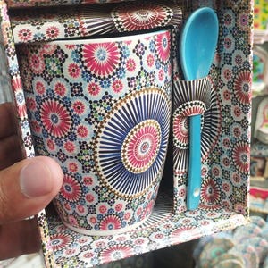 Puede incluir: Una taza de cerámica decorativa con una cuchara a juego, presentada en una caja estampada. La taza presenta un vibrante diseño floral en rosa, azul y blanco. La cuchara es de color turquesa. La caja y la tapa tienen un patrón coordinado.