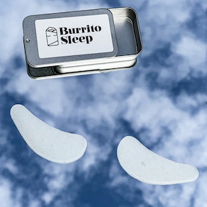 Puede incluir: Una lata plateada con las palabras "Burrito Sleep" impresas en la tapa. Dos parches para ojos blancos en forma de media luna están sobre un fondo nublado azul y blanco.