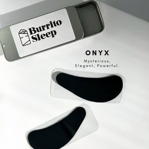 May include: A silver metal case with the text "Burrito Sleep" and a graphic of a sleeping burrito. Two black, curved eye masks are displayed below, with the word "ONYX" and the description "Mysterious, Elegant, Powerful."