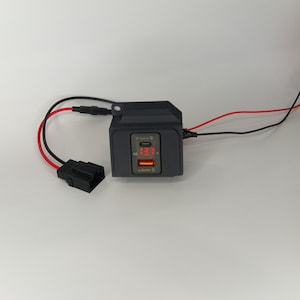 May include: A black car charger with two USB ports and a digital display showing 13.7 volts. The charger has red and black wires attached.