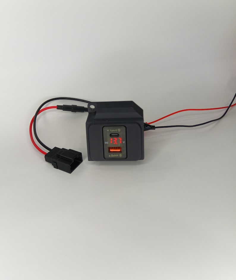 May include: A black car charger with two USB ports and a digital display showing 13.7 volts. The charger has red and black wires attached.
