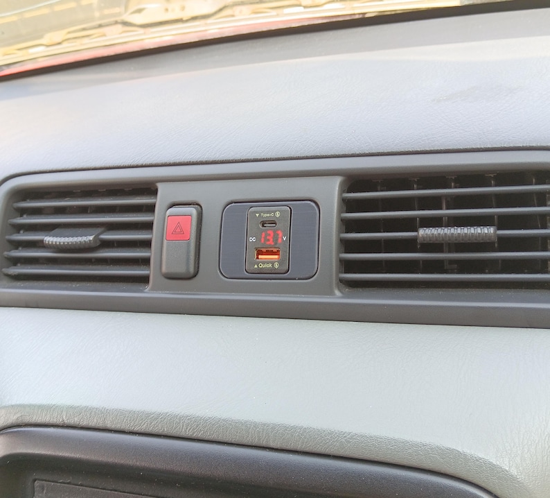 May include: A close-up of a car's dashboard with a black and red emergency button and a digital display showing 13.1V. The display is part of a car charger with a USB port and a Quick Charge logo.