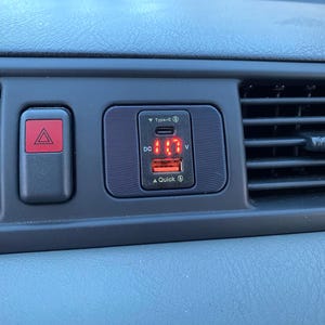 May include: A black car charger with a red digital display showing "11.7" and the text "DC" and "V". The charger has a Type-C port and a Quick Charge port.