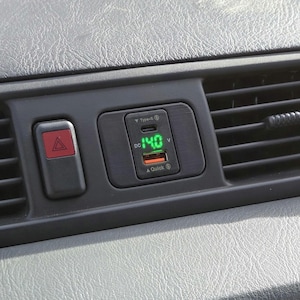 Puede incluir: Un cargador de coche negro con una pantalla digital verde que muestra "14,0 V". El cargador tiene un puerto USB-C y un logotipo de carga rápida.