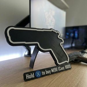 Puede incluir: Un organizador de escritorio negro y blanco con forma de pistola con un soporte. El organizador tiene un contorno blanco y el texto "Hold X to buy M1911 (Cast: 1000)". Una pistola real es visible en el fondo.