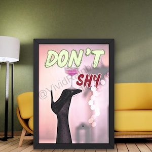 May include: Framed art print featuring a black high heel shoe and a glass of pink liquid. The words "DON'T SHY" are displayed in green and red. The image is set against a pink and white background.