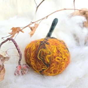 May include: A handmade felt pumpkin in shades of orange, yellow, and brown, with a green stem. The pumpkin is set against a backdrop of white fluffy material, with dried leaves and twigs adding a natural touch.