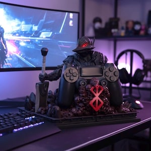 May include: A black controller stand featuring a warrior figure holding a sword, with a red symbol and skull details. The stand is designed to hold a game controller, with a keyboard and monitor in the background.