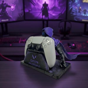 May include: A white and black PlayStation 5 controller sits on a black stand with the text "OMEN VALORANT". A purple figure is positioned behind the controller. Computer monitors displaying video game graphics are in the background.