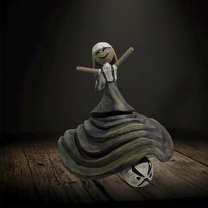 May include: A whimsical wooden figurine of a dancing figure with arms outstretched. The figure wears a white and black striped top, a long gray skirt, and a white hat. The figurine is perched atop a ball with a black and white face.