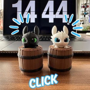 May include: Two 3D-printed dragon figurines, one black with green eyes and the other white with blue eyes, sit atop small brown barrel-shaped bases. The word "CLICK" is displayed in a blue and white bubble font at the bottom of the image.