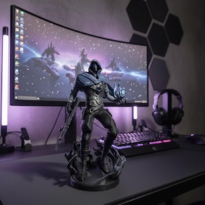 May include: A detailed figurine of a character from a video game, standing on a base. The figure is predominantly black and gray, with blue accents. It is holding a weapon and a glowing blue orb. The background includes a computer monitor and gaming accessories.