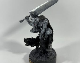 Puede incluir: Una figura de plata y negro de Guts de la serie de manga Berserk. Él está sosteniendo una espada grande y lleva armadura. La figura está de pie sobre una base negra con la palabra "Berserk" escrita en ella.