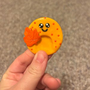 Peut inclure: Un charm circulaire en argile jaune arborant un visage souriant avec des yeux et une bouche noirs. Le charm est décoré de petits points orange et d'une feuille d'érable orange texturée fixée sur le côté gauche, créant un accessoire automnal.