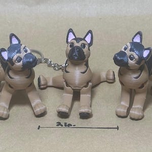 May include: Three German Shepherd keychains. Each keychain is a miniature, tan-colored dog with black and gray markings. The dogs have a detailed design, including ears, eyes, and a nose. The keychains are connected by a chain and a keyring. The image includes a 2-inch measurement.