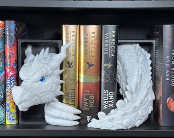 Pode incluir: Suportes de livros de dragão branco com olhos azuis, segurando uma coleção de livros em uma prateleira preta. Os livros têm capas douradas, pretas e coloridas. Os títulos incluem "Onyx Storm" e "Iron Flame."