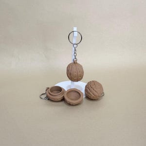 May include: A collection of walnut-shaped keychains and a walnut-shaped container. One keychain is attached to a metal ring and hangs from a white stand. The other keychains and container are on a neutral background. The keychains and container are brown.