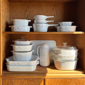 May include: A set of white casserole dishes with a blue floral pattern. The dishes are arranged on a wooden shelf in a kitchen cabinet.