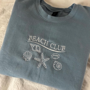 May include: A light blue sweatshirt with the text "Beach Club" embroidered in white. The design also includes embroidered white seashells and a starfish.