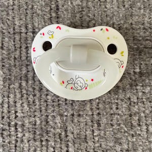 以下が含まれることがあります： サルと鳥のデザインが施された白いおしゃぶり。おしゃぶりは白いハンドルと側面に2つの小さな穴があります。