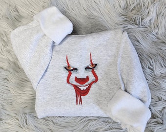 May include: A light gray sweatshirt featuring an embroidered image of a clown face with red accents. The clown has orange eyes, a red nose, and a wide, sinister smile. The sleeves are folded, and the background is a fluffy, light gray surface.