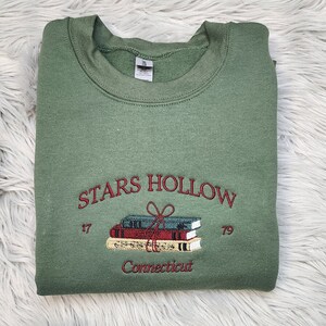 K&ouml;nnte beinhalten: Salbeigr&uuml;nes Sweatshirt mit gesticktem Text "STARS HOLLOW" &uuml;ber einem Stapel B&uuml;cher, die mit einer Schleife zusammengebunden sind. Die Texte "17" und "79" flankieren die B&uuml;cher, darunter "Connecticut". Das Sweatshirt ist gefaltet.