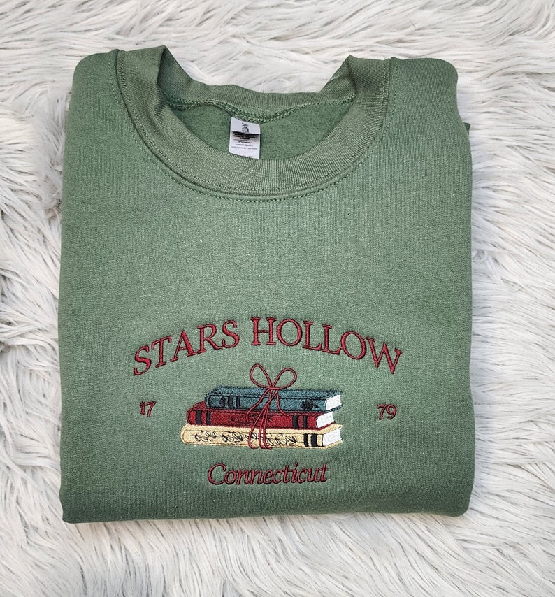 K&ouml;nnte beinhalten: Salbeigr&uuml;nes Sweatshirt mit gesticktem Text "STARS HOLLOW" &uuml;ber einem Stapel B&uuml;cher, die mit einer Schleife zusammengebunden sind. Die Texte "17" und "79" flankieren die B&uuml;cher, darunter "Connecticut". Das Sweatshirt ist gefaltet.