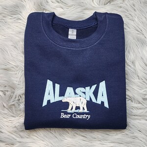 K&ouml;nnte beinhalten: Marineblaues Sweatshirt mit dem Wort "ALASKA" in Hellblau, einer Eisb&auml;rgrafik und dem Text "Bear Country". Das Sweatshirt ist gefaltet und scheint aus einem weichen Material zu sein.