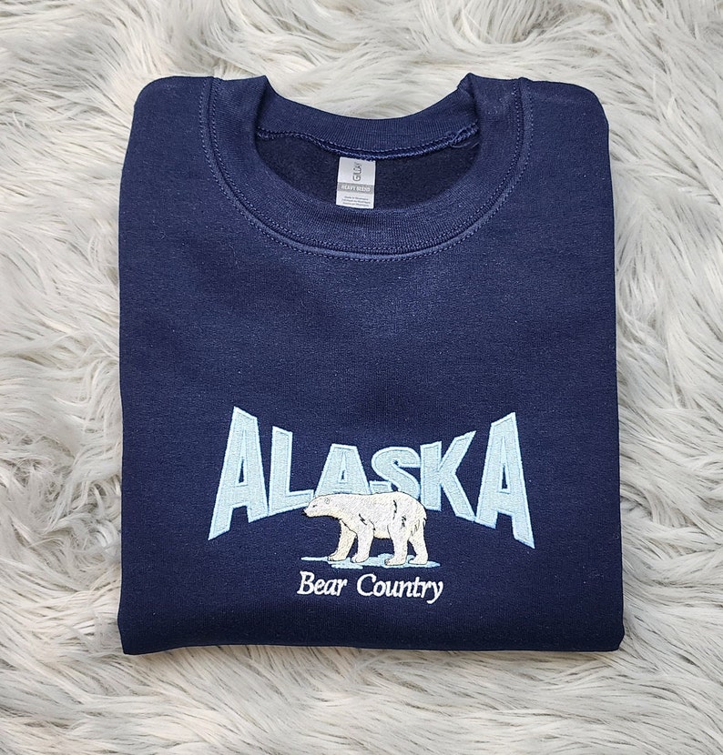 K&ouml;nnte beinhalten: Marineblaues Sweatshirt mit dem Wort "ALASKA" in Hellblau, einer Eisb&auml;rgrafik und dem Text "Bear Country". Das Sweatshirt ist gefaltet und scheint aus einem weichen Material zu sein.
