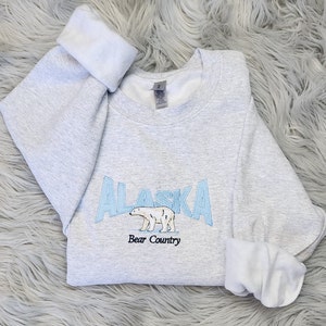 K&ouml;nnte beinhalten: Hellgraues Sweatshirt mit dem in Hellblau gestickten Wort "ALASKA", einer Eisb&auml;renillustration und dem Text "Bear Country". Die &Auml;rmel sind umgeschlagen und zeigen ein wei&szlig;es Innenfutter.