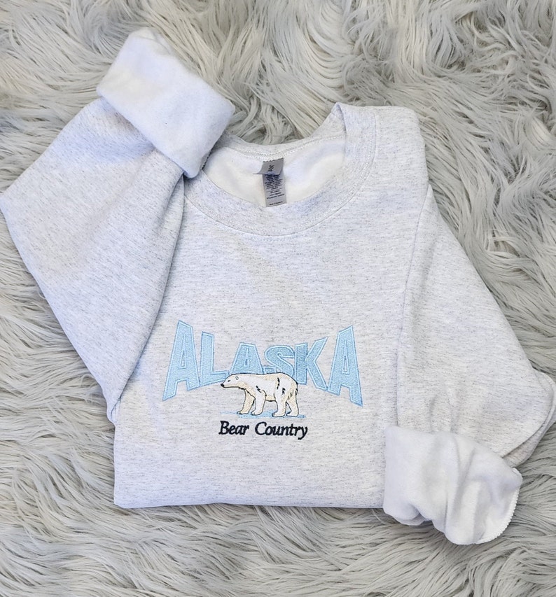 K&ouml;nnte beinhalten: Hellgraues Sweatshirt mit dem in Hellblau gestickten Wort "ALASKA", einer Eisb&auml;renillustration und dem Text "Bear Country". Die &Auml;rmel sind umgeschlagen und zeigen ein wei&szlig;es Innenfutter.