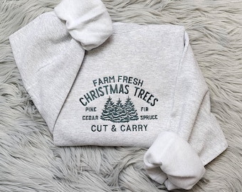 May include: A heather gray sweatshirt with the text "FARM FRESH CHRISTMAS TREES" in dark green. The design includes illustrations of pine trees and the words "PINE, CEDAR, FIR, SPRUCE, CUT & CARRY". The sweatshirt has a hood and long sleeves.