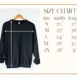 K&ouml;nnte beinhalten: Ein schwarzes Sweatshirt h&auml;ngt an einem Holzb&uuml;gel, mit einer Gr&ouml;&szlig;entabelle rechts. Die Gr&ouml;&szlig;entabelle zeigt die Breiten- und L&auml;ngenma&szlig;e in Zoll f&uuml;r die Gr&ouml;&szlig;en S, M, L, XL und 2X.