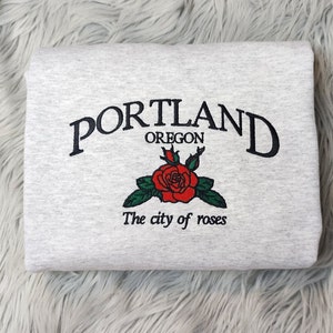 Könnte beinhalten: Ein gefaltetes hellgraues Sweatshirt mit dem gestickten Text "PORTLAND OREGON" über einer roten Rose mit grünen Blättern. Unter der Rose steht der Text "The city of roses".