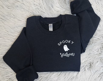 May include: Black crewneck sweatshirt with the words "Spooky Season" and a white ghost graphic. The sweatshirt is folded, with the sleeves cuffed. The text and graphic are in white.