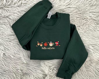 May include: A dark green sweatshirt with embroidered autumn-themed designs, including acorns, leaves, a coffee cup, and a mushroom. The words "hello autumn" are embroidered below the designs. The sweatshirt is displayed on a fluffy, light-colored surface.