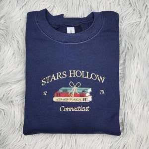 K&ouml;nnte beinhalten: Marineblaues Sweatshirt mit gesticktem Text und Grafik. Der Text lautet "STARS HOLLOW" &uuml;ber einem Stapel B&uuml;cher, die mit einer Schleife gebunden sind, mit "17" und "79" auf beiden Seiten. Unter den B&uuml;chern steht "Connecticut".