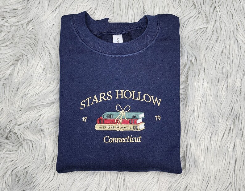 K&ouml;nnte beinhalten: Marineblaues Sweatshirt mit gesticktem Text und Grafik. Der Text lautet "STARS HOLLOW" &uuml;ber einem Stapel B&uuml;cher, die mit einer Schleife gebunden sind, mit "17" und "79" auf beiden Seiten. Unter den B&uuml;chern steht "Connecticut".
