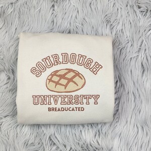 May include: Cream-colored sweatshirt with the text "SOURDOUGH UNIVERSITY" in brown lettering, along with a graphic of a loaf of bread. The word "BREADUCATED" is printed below the university name.